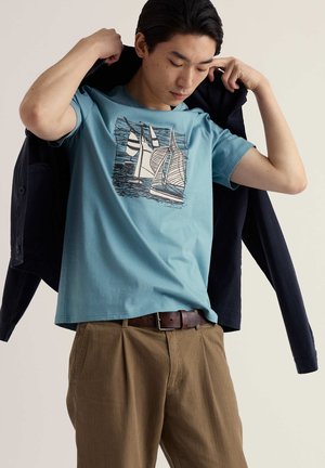 Mees, kes kannab sinist T-särki purjekarajooniga, paneb selga tumeda jopi, mille juurde kuuluvad pruunid püksid ja tumevöö.