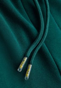 Tejido verde con una textura suave presenta dos cordones trenzados que terminan en puntas de plástico transparente, etiquetadas como "LEVEL UP" en amarillo.