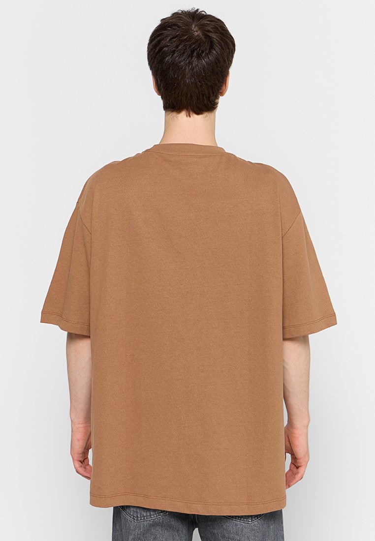 Mężczyzna z krótkimi ciemnymi włosami, ubrany w za dużą brązową koszulkę i ciemne dżinsy, widziany z tyłu na jednolitym jasnym tle.