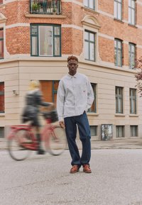 Άνδρας με ριγέ πουκάμισο και τζιν στέκεται σε γωνία δρόμου μπροστά από κτίριο με τούβλα, ενώ ένας θαμπός ποδηλάτης περνάει δίπλα.