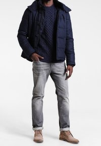 Granatowa kurtka puchowa na granatowym swetrze w warkocz, połączona z jasnoszarymi dżinsami i beżowymi botkami za kostkę. Czyste linie, zdefiniowany kształt.