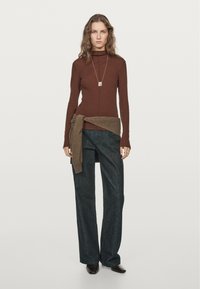 Brown long-sleeve turtleneck sweater with a textured finish, paired with loose dark blue denim pants and a tied brown sweater at the waist.