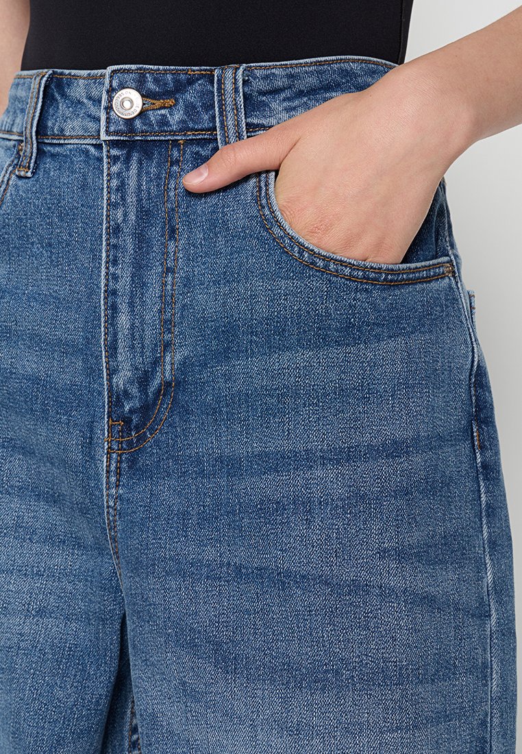Jean en denim bleu taille haute avec une coupe droite. Les caractéristiques incluent une poche avant, des coutures contrastantes en brun et une fermeture à bouton en métal.
