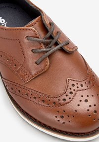 Brown leather dress shoe featuring intricate brogue detailing, rounded toe, and contrasting dark laces. Smooth texture with a white sole.