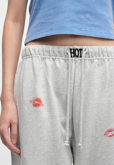 Vidukļa apģērbs viegli pelēkās sporta biksēs ar sarkanu lūpu krāsas skūpstu nospiedumiem un uzrakstu "HOT" uz jostas, kopā ar zilu īsu topi.