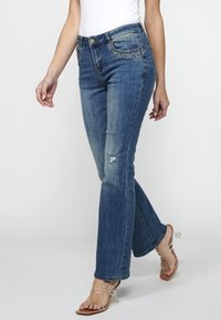 Vaqueros de mezclilla azul acampanados con detalles desgastados en las rodillas, bolsillos adornados con strass y cintura ajustada.