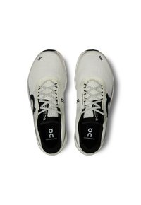 Witte sportieve schoenen met een gestructureerde bovenkant, zwart logo, reflecterende accenten en een ronde zool. De veters zijn wit met een patroon.