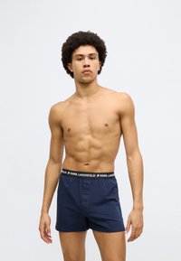 Μπόξερ από βαμβάκι σε ναυτικό μπλε με μαύρη ζώνη μέσης με λογότυπο, διαθέτει κουμπωτό εμπρός και ομαλή υφή. Το μοντέλο παρουσιάζει ουδέτερη στάση.