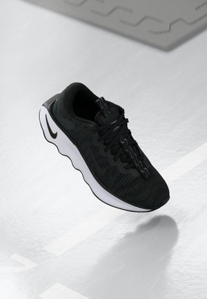 Chaussure de sport noire avec semelle blanche ondulée flottant au-dessus d’un sol blanc avec ombre, montrant les détails latéraux et supérieurs, y compris les lacets et le logo.