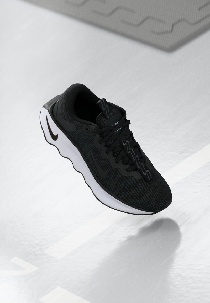 Chaussure de sport noire avec semelle blanche ondulée flottant au-dessus d’un sol blanc avec ombre, montrant les détails latéraux et supérieurs, y compris les lacets et le logo.