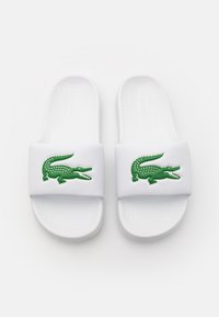 Des sandales blanches à enfiler, avec un semelle texturée et un logo crocodile vert bien en vue sur la sangle.