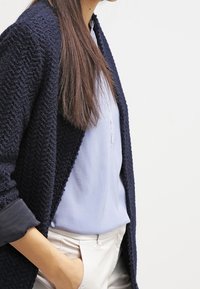 Cardigan lavorato a maglia di colore navy con un motivo testurizzato, indossato sopra una blusa azzurro chiaro. Presenta maniche lunghe e una vestibilità rilassata.