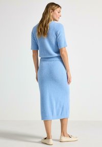 Kvinna stående med ryggen vänd, iklädd en ljusblå mjuk kortärmad tröja och matchande midikjol med krämfärgade loafers.