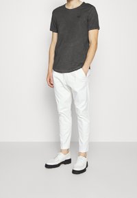 Uomo che indossa una maglietta a maniche corte grigio carbone, pantaloni bianchi slim-fit e scarpe bianche con lacci e suola nera, in piedi con le mani in tasca.