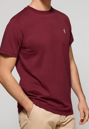 Homme portant un t-shirt bordeaux ajusté à manches courtes avec un petit logo blanc brodé sur le côté gauche de la poitrine et un pantalon beige.