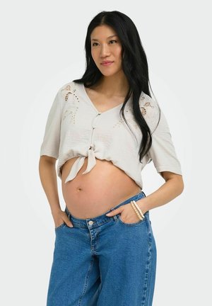 Gravid kvinne med langt svart hår iført en lys cropped bluse og blå jeans, hendene i lommer, ser litt til siden.