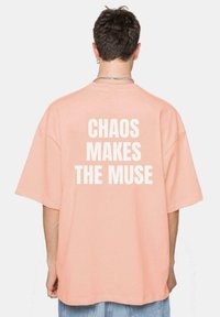 Camiseta de melocotón de gran tamaño con mangas cortas que presenta un audaz texto blanco en la parte posterior que dice "EL CAOS HACE A LA MUSA." Textura suave.