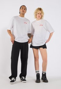 Dva lidé mají na sobě bílé oversized trička s červeným textem, které doplňují černé kalhoty a šortky. Pozadí je jednobarevné bílé.