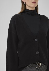 Cardigan negru, din tricot moale, cu decolteu adânc în V, butoni frontali și mâneci largi, purtat peste un pulover negru cu guler înalt.