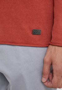 Röd stickad tröja med texturerad yta, svart logoplack vid fållen och kontrasterande grått tyg nedanför.