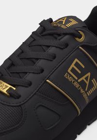 Μαύρο αθλητικό παπούτσι με μαύρα κορδόνια, χρυσές τρύπες για κορδόνια και χρυσό λογότυπο Emporio Armani EA7 στη γλώσσα και στην πλευρική επιφάνεια.