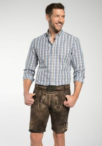 Hartige bruine lederhosen met decoratieve stiksels, gecombineerd met een blauw-wit geruite button-up shirt. Model glimlacht, handen in de zakken.