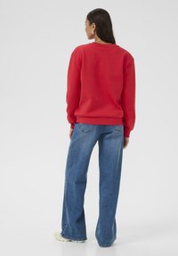 Czerwony sweter z okrągłym dekoltem, luźny krój, ściągacze na rękawach, zestawiony z jasnobłękitnymi spodniami typu straight-leg i beżowymi trampkami. Gładka faktura.