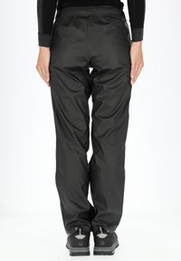 Zwarte waterdichte broek met een verstelbare elastische tailleband, slank ontwerp en elastische manchetten. Ggedragen met zwarte wandelschoenen.