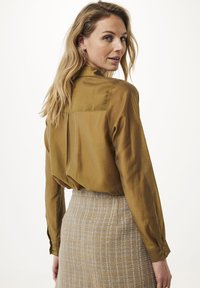 Satinbluse in Olivgrün mit Rückenfalte, langen Ärmeln und geknöpften Manschetten, kombiniert mit einem strukturierten, grau-gelben gewebten Rock.