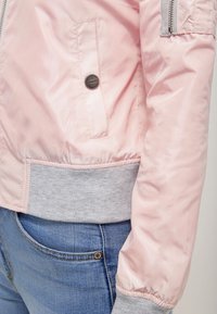 Gros plan sur une veste bomber rose avec une taille et des poignets côtelés gris, portée par-dessus un jean en denim bleu.