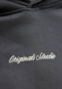 Tessuto del felpa grigio con una texture liscia. Presenta "Originals Studio" ricamato in carattere corsivo bianco sul davanti. Design semplice e minimalista.