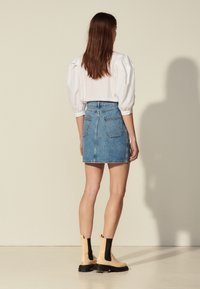 Mini-jupe en jean bleu clair avec poches arrière, associée à un chemisier blanc à manches bouffantes et des bottines beiges avec des accents noirs.