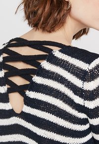 Pruhovaný pletený svetr v námořnické modři a bílé barvě s designem překřížených pásků na zádech a otevřenými štěrbinami. Měkká textura, volný střih a tříčtvrteční rukávy.
