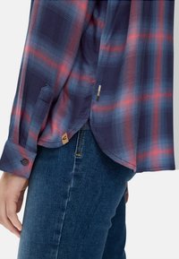 Plaid shirt in navy, red, and light blue. Long sleeves with buttoned cuffs and a rounded hem. Features logo tag at the side. Denim jeans visible.