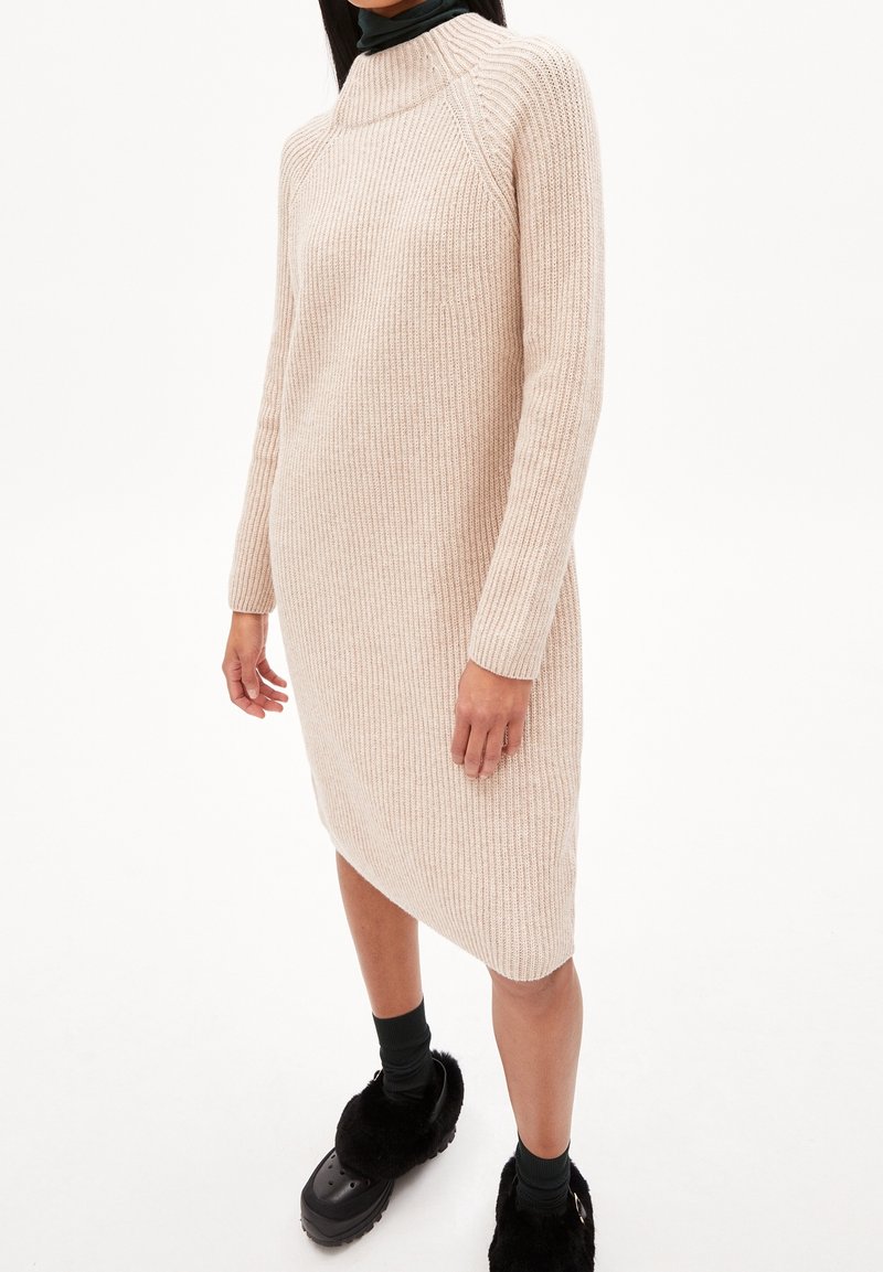 Femme portant une robe en maille côtelée beige à manches longues avec un ourlet asymétrique et des chaussures noires fourrées à enfiler.
