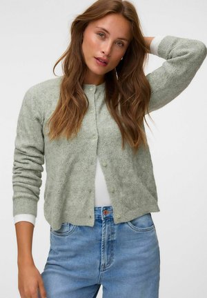 Jeune femme aux longs cheveux bruns porte un cardigan tricoté vert clair sur un haut blanc et un jean bleu, posant avec un bras levé derrière la tête.