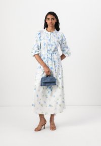 Vestido midi floral em branco e azul com detalhes em verde, apresentando uma cintura com cinto, mangas franzidas e colarinho redondo; combinado com uma mala azul.