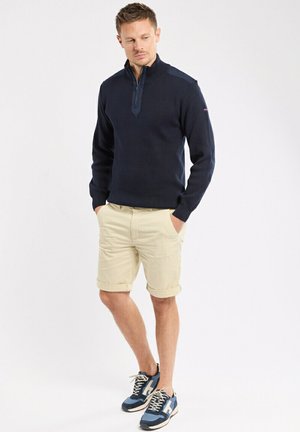 Uomo in piedi con le mani nelle tasche che indossa un maglione blu navy con mezza cerniera, pantaloncini beige e scarpe da ginnastica blu su sfondo bianco.