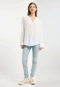 Blusa bianca leggera a maniche lunghe con scollo a V e bottoni, abbinata a jeans skinny azzurri e sneaker alte beige.