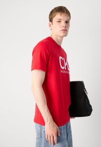 Raudona medvilninė marškinė su dideliu baltu "CK" logotipu, derinama su šviesiai mėlynomis džinsinėmis kelnėmis ir juoda rankine su sidabriniu užtrauktuku.