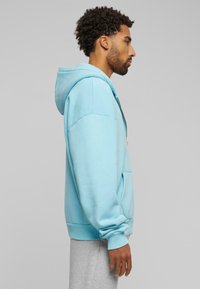 Világoskék kapucnis pulóver, amely pamutból készült, elülső zsebbel, húzózsinórral a kapucnin és bordás mandzsettákkal. Szürke melegítőnadrág fölött viselve, oldalról látható.