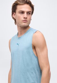 Νεαρός άνδρας με ανοιχτό καστανά μαλλιά, που φοράει ένα ανοιχτό μπλε αμάνικο μπλουζάκι Puma, κοιτάζει στο πλάι πάνω σε απλό φόντο.