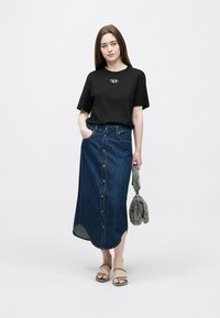 Juoda trumpomis rankovėmis marškinėliai su logotipu, mėlyna džinsinė sijonas su sagomis priekyje, lygios basutės, laikanti rankinę su raštu. Neutralus fonas.