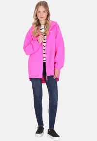 Világos rózsaszín vízálló dzseki kapucnival, cipzáras elülső résszel, elasztikus mandzsettákkal, csíkos ing fölött viselve. Sötét farmerral és fekete sportcipővel párosítva.