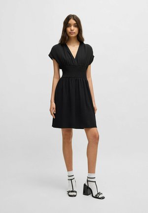 Black dress with a deep V-neck, short sleeves, and a smocked waist. A textured fabric falls to mid-thigh, paired with heeled sandals and socks.