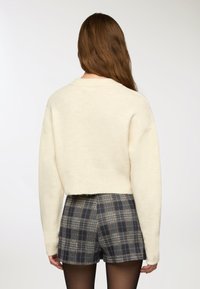 Maglione corto crema con maniche lunghe, tessuto a maglia. Abbinato a pantaloni corti a quadri nei colori blu navy, grigio e bianco.
