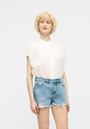 Nainen, jolla on kiharat vaaleat hiukset ja päällään teksturoitu valkoinen hihaton pusero sekä vaaleansiniset kulutetut farkkushortsit, seisoo yksinkertaista taustaa vasten.