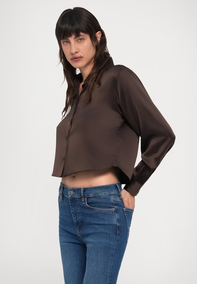 Hnedá saténová krátka košeľa s dlhými rukávmi a zapínaním na gombíky, kombinovaná s modrými džínsami z denimu so slabým opotrebovaním.