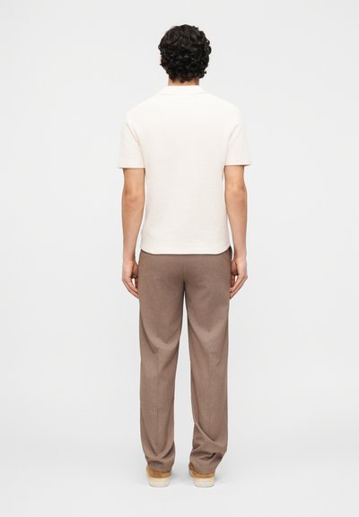 Άνδρας με την πλάτη, φοράει λευκό κοντομάνικο πόλο πουκάμισο, καφέ παντελόνια και μπεζ παπούτσια σε απλό φόντο.