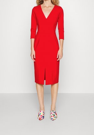 Femme portant une robe rouge à col en V, longueur genou, manches trois quarts, et des chaussures pointues colorées à pois, debout devant un fond blanc uni.